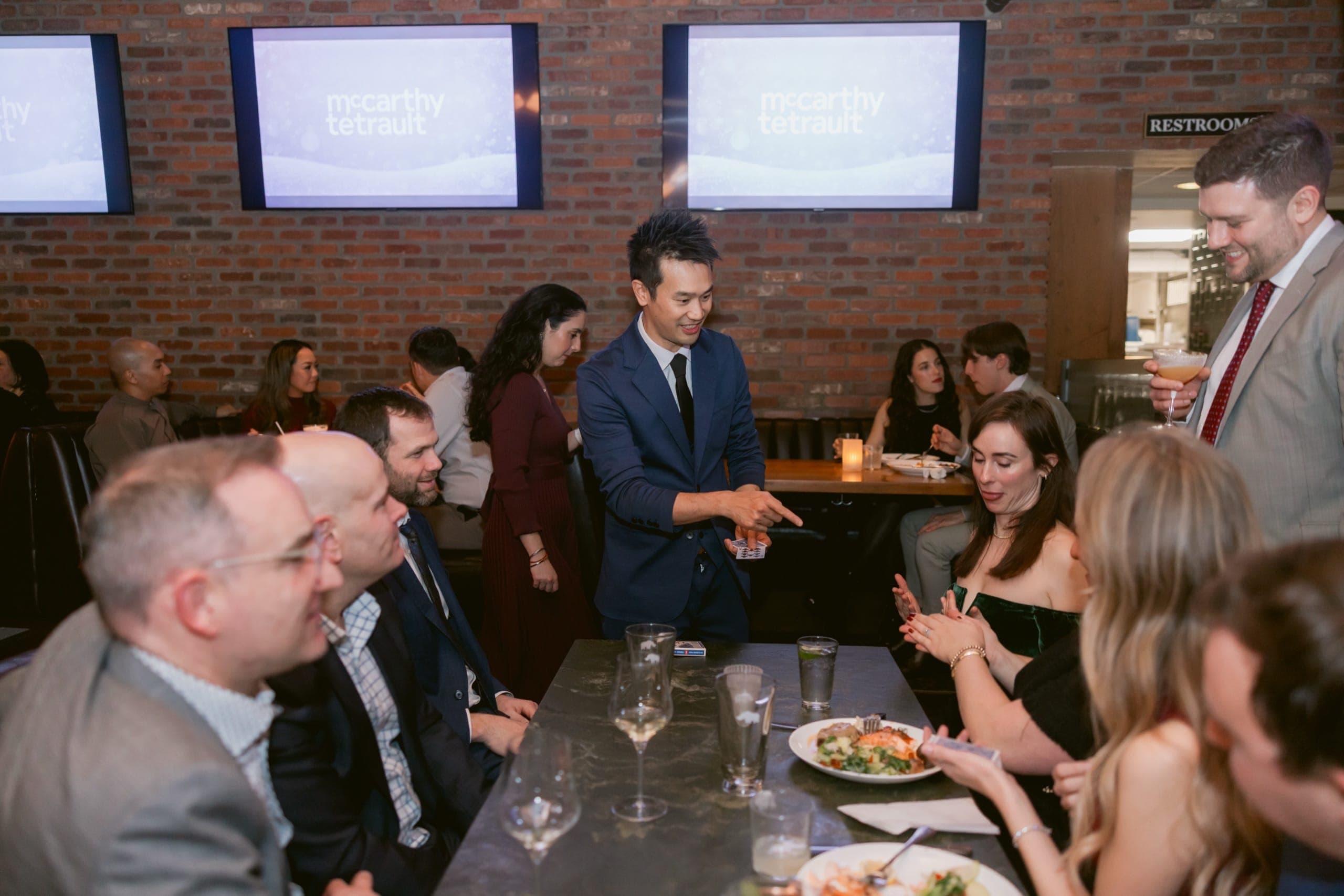 Kel performing close-up card magic for McCarthy Tétrault guests at their Vancouver holiday party, with the firm's logo visible on screens behind him